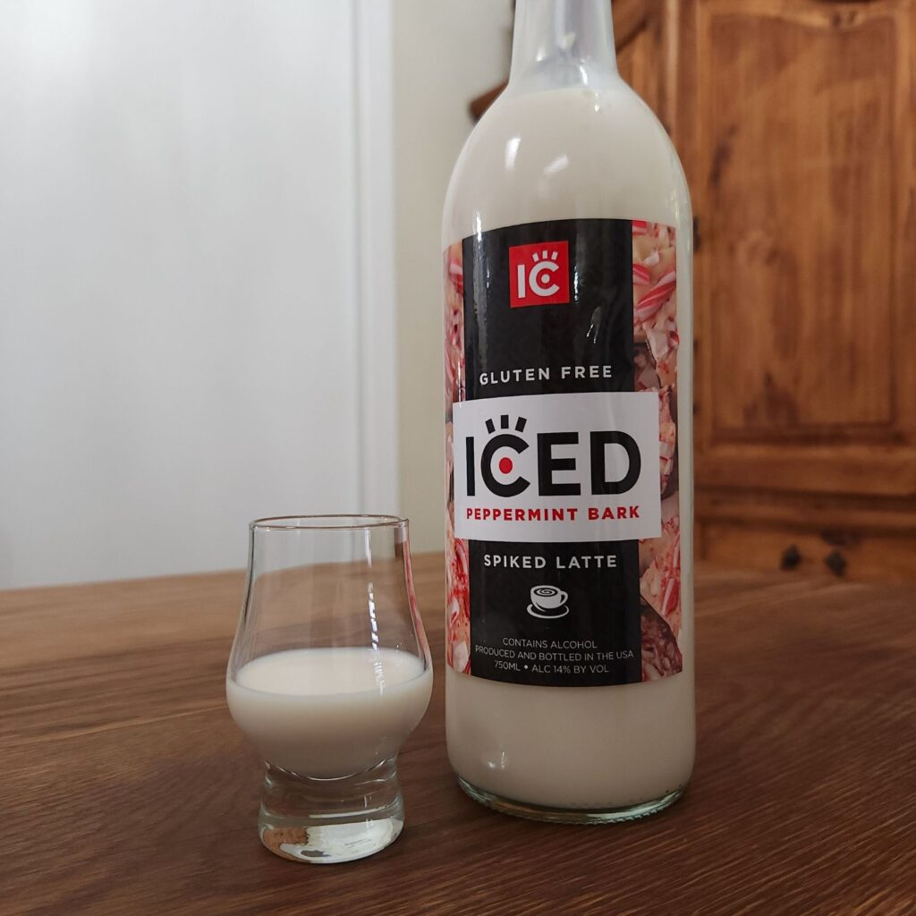 Bottle of IC Iced Peppermint Bark Spiked Latte with only the bottom two thirds of the bottle showing, next to a small snifter glass with creamy beige white liquid, both sitting on a wooden table