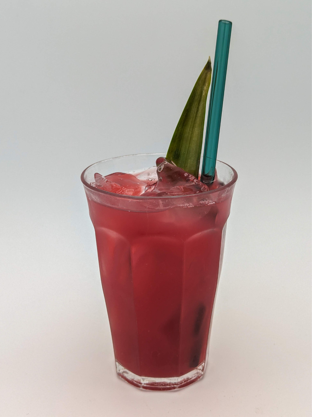 Ruby red colored liquid in a old fashioned glass with a green straw and a pineapple leaf garnish