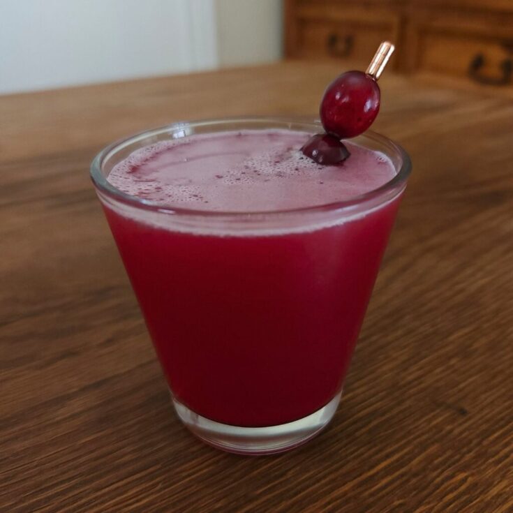 Small angled glass filled with deep red liquid, with a rose gold cocktail pick with fresh cranberries sticking out of the glass at a diagonal angle