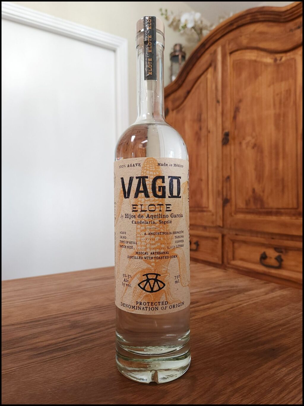 Bottle of Mezcal Vago Elote sitting on a wooden table, in front of a mixed white and wooden background