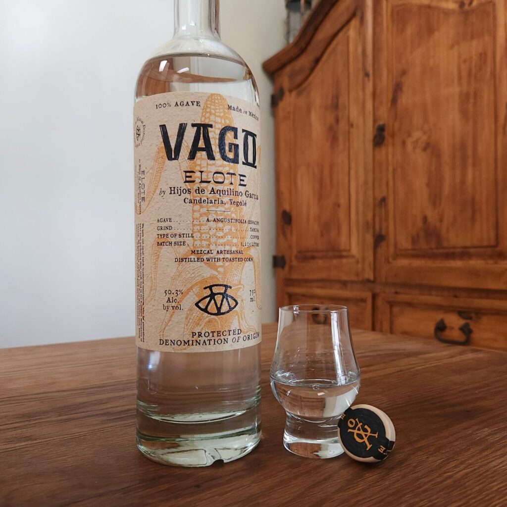 Bottle of Mezcal Vago Elote with the lower two thirds showing, next to the cork top and a small snifter glass with clear liquid, all sitting on a wooden table