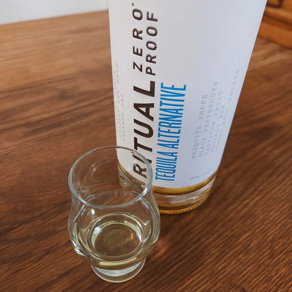 Bottle of Ritual Zero Proof Tequila alternative with only the label showing, next to a small snifter glass with light golden liquid, both sitting on a wooden table