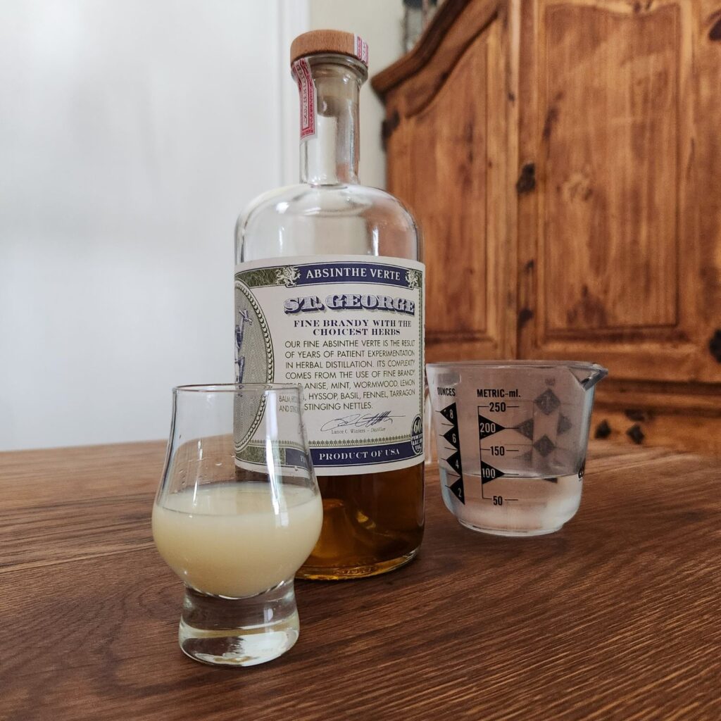 Half full bottle of St. George Absinthe Verte next to a small measuring glass with clear liquid, and a snifter glass with cloudy light yellow liquid, all sitting on a wooden table