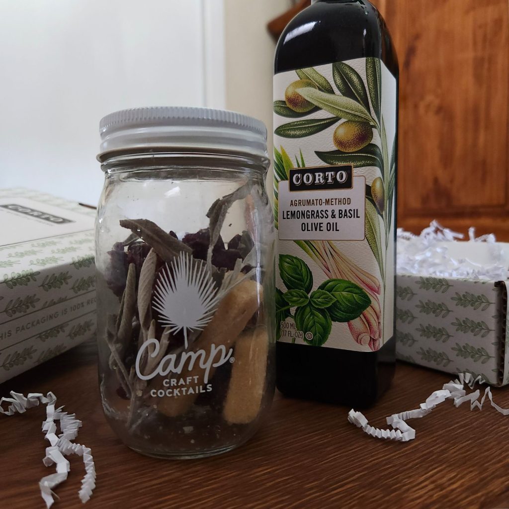 Jar filled with dehydrated botanicals next to a bottle of olive oil with only the label showing, both sitting on a wooden table in front of an open box in white and light green, with white packing materials peeking above the edge