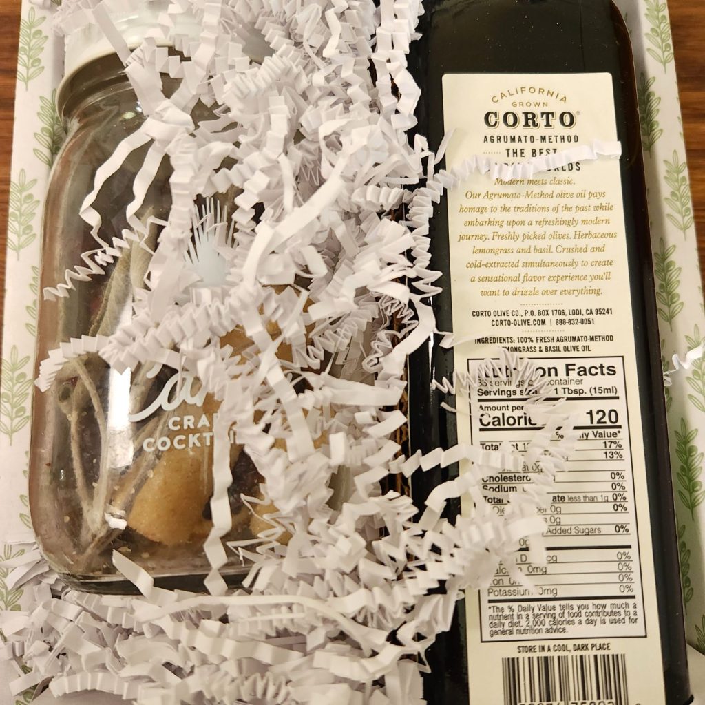 Cardboard box in white and light green, filled with a jar of dehydrated botanicals and a bottle of olive oil with the back label showing. White strips of paper act as packing material between and partially in front of the contents.