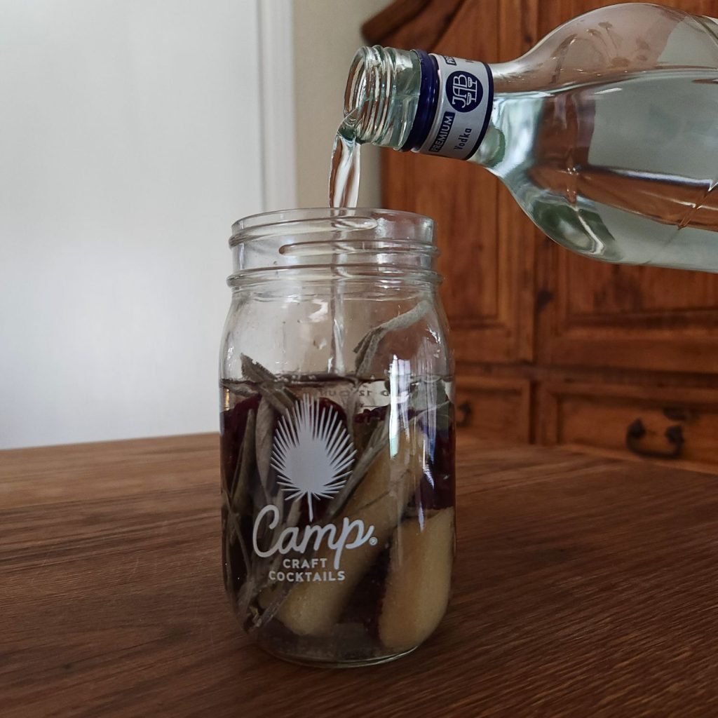 Open jar filled with dehydrated botanicals, with a bottle of vodka with only the top part showing pouring clear liquid into the jar