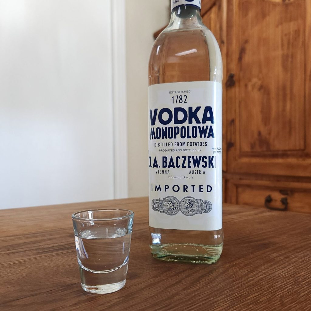 Bottle of Monopolowa Vodka with the bottom 90 percent of the bottle showing, next to a shot glass with clear liquid, both sitting on a wooden table