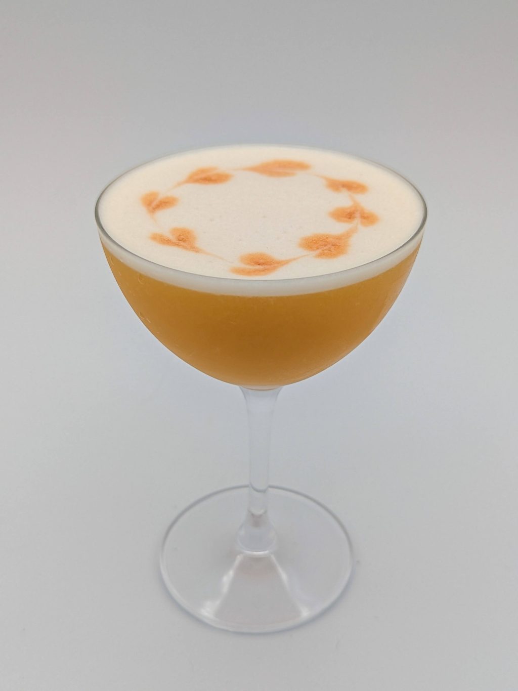 Golden liquid with a white foam top with orange angostura bitters as garnish in a coup glass