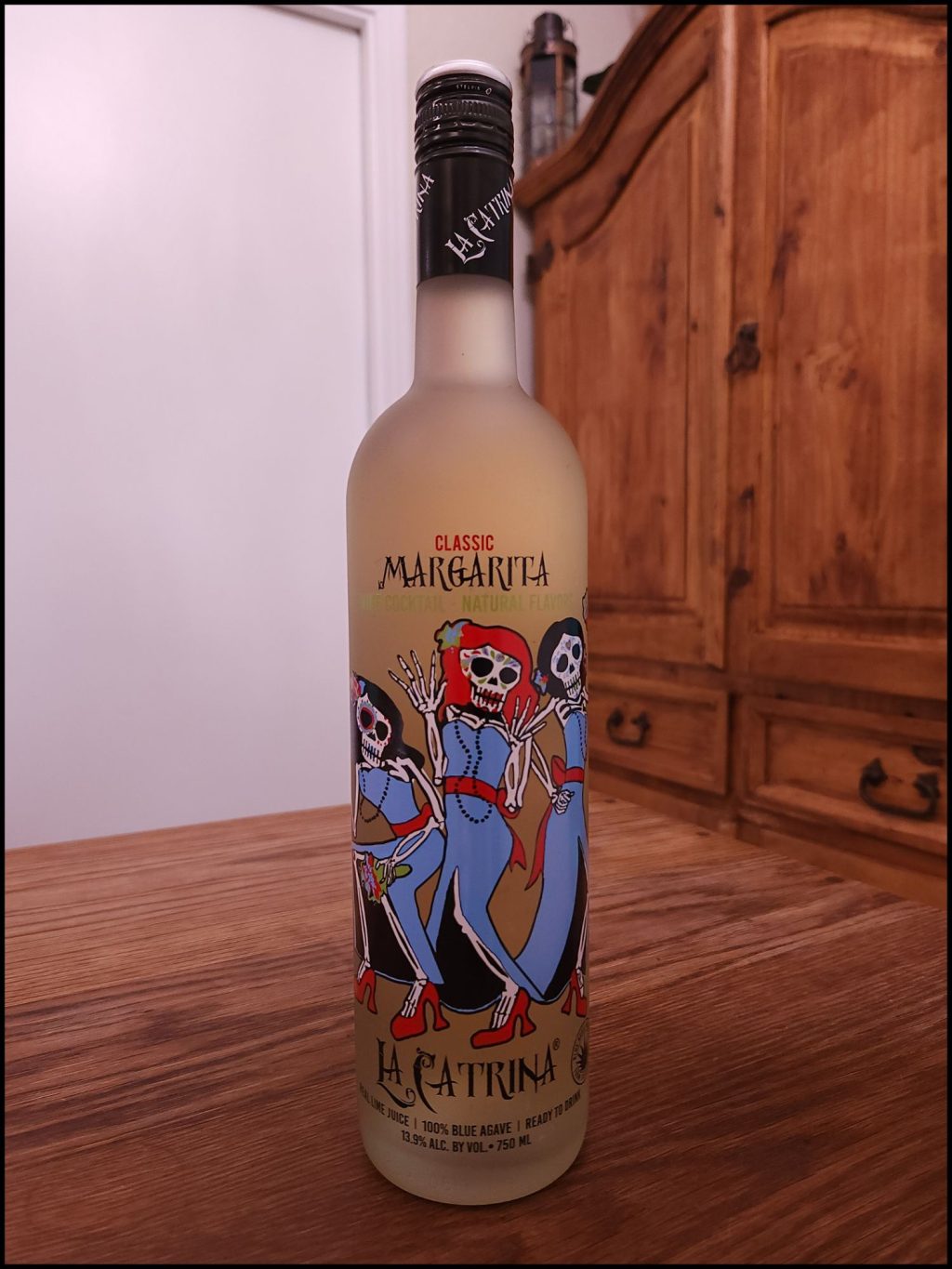 Bottle of La Catrina Classic Margarita with frosted glass and a light blue, red, and black label design, sitting on a wooden table