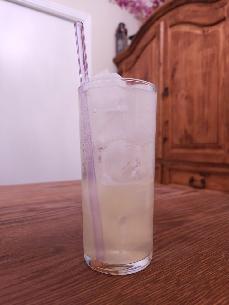 Highball glass with ice, light yellow liquid, and a light purple glass straw, sitting on a wooden table