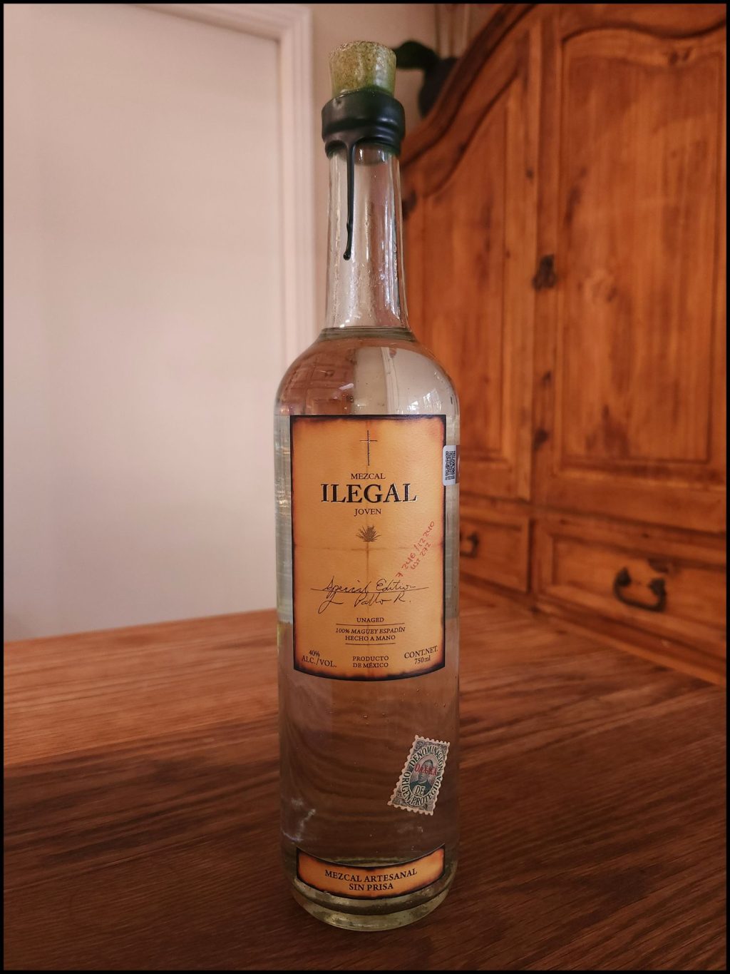 bottle of Mezcal Ilegal Joven with clear glass and a yellow beige label design, sitting on a wooden table