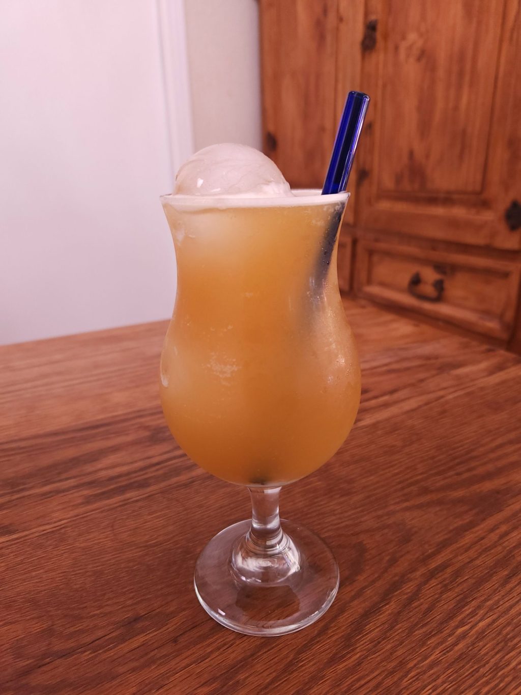 Hurricane glass with ice, golden beige liquid, and a cobalt blue glass straw