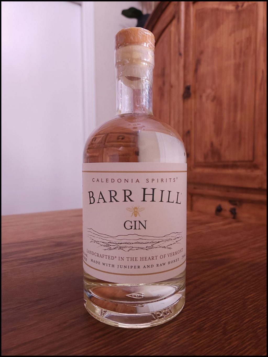 Bottle of Barr Hill Small Batch Gin with clear liquid and an off-white label design, sitting on a wooden table