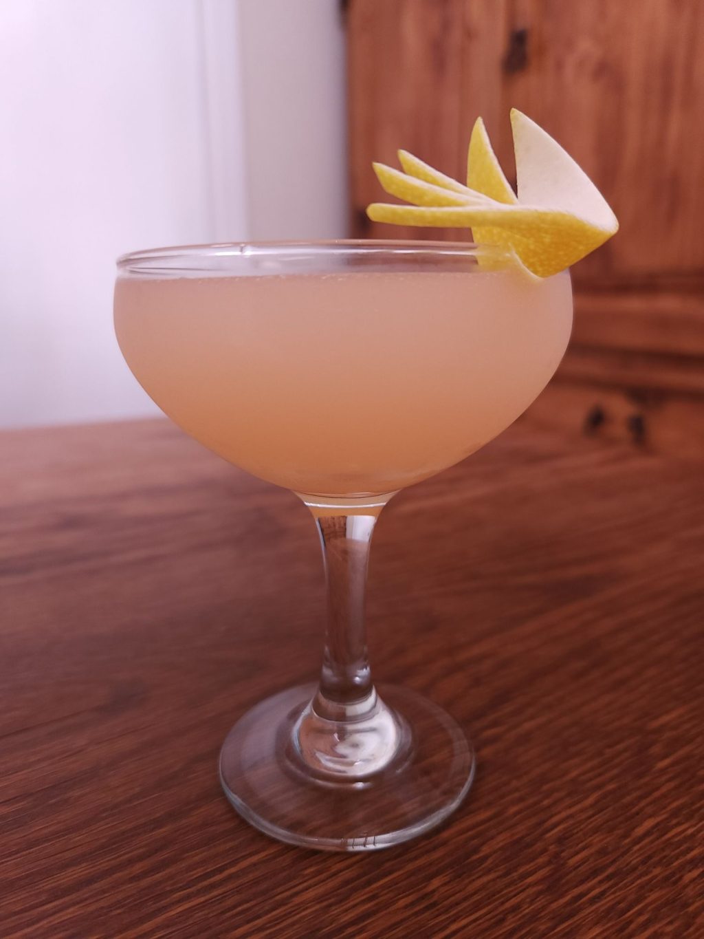 Coupe glass with light orange pink liquid and an artistic lemon peel garnish, sitting on a wooden table