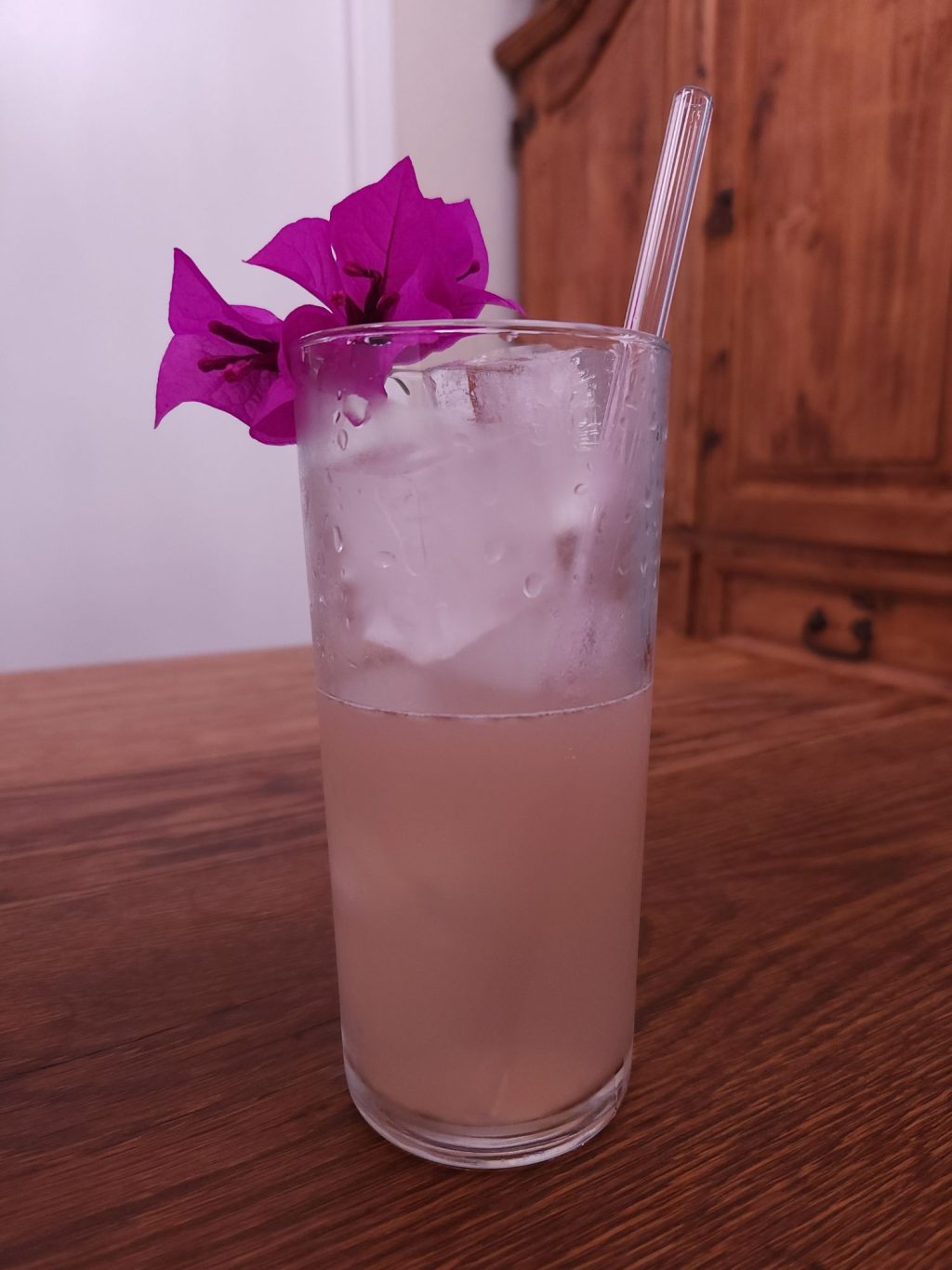 Tall collins glass with ice and light pink liquid, a light pink glass straw, and a bright purple pink bougainvillea blossom garnish