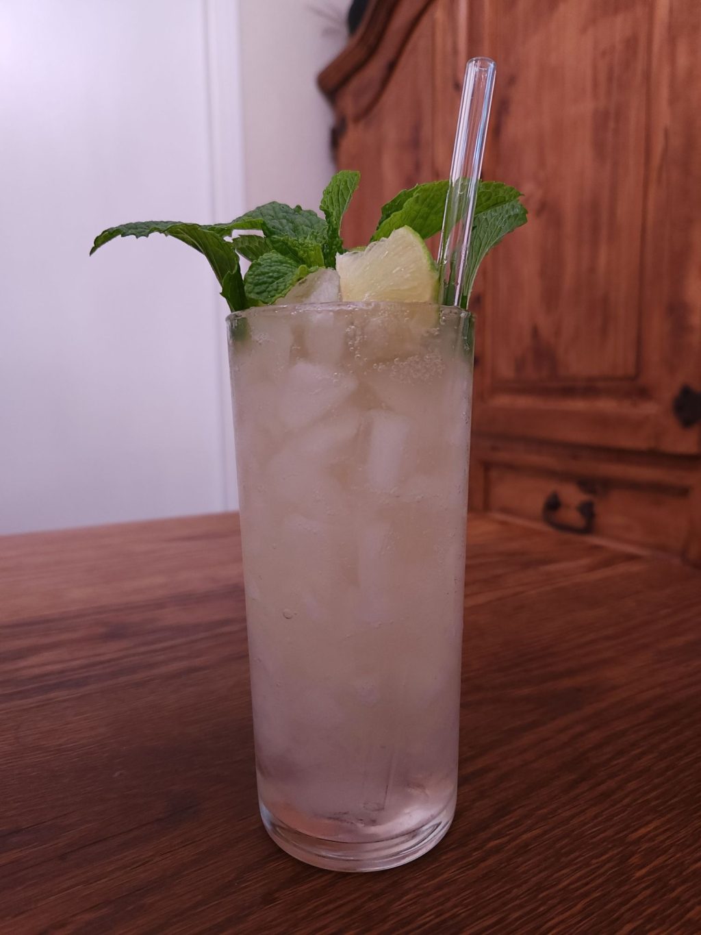 Highball glass with ice, light beige liquid, a glass straw, and a mint and lime wedge garnish