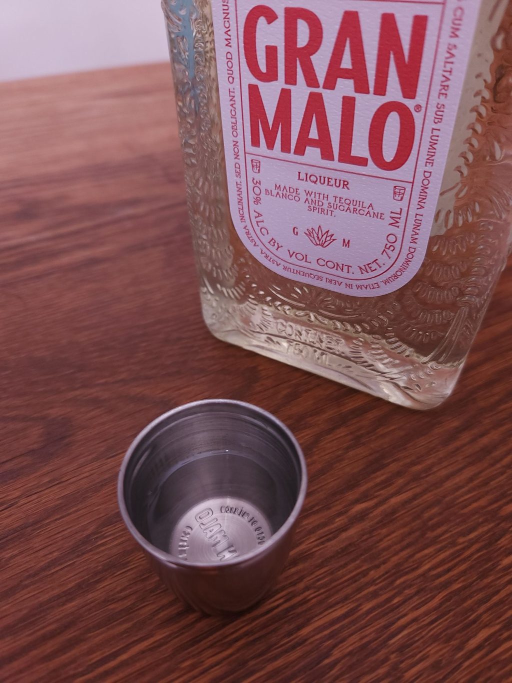 metal shot glass with yellowish clear liquid, next to a bottle of Gran Malo Tamarind Liqueur with the bottom part of the red and white label showing