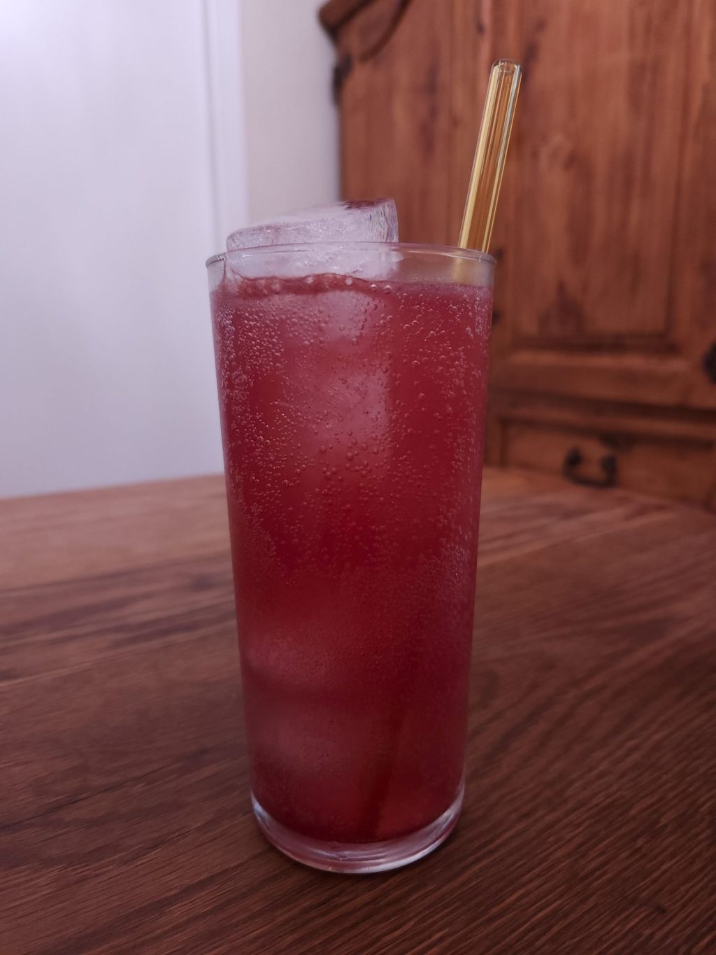 Tall glass with ice and deep red colored carbonated liquid and a yellow glass straw, sitting on a wooden table