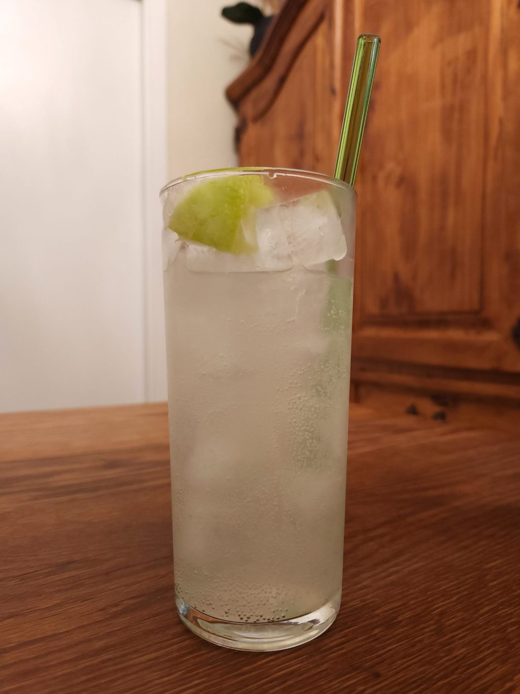 Tall glass with ice and very light pink carbonated liquid, a green glass straw, and a squeezed out lime wedge garnish