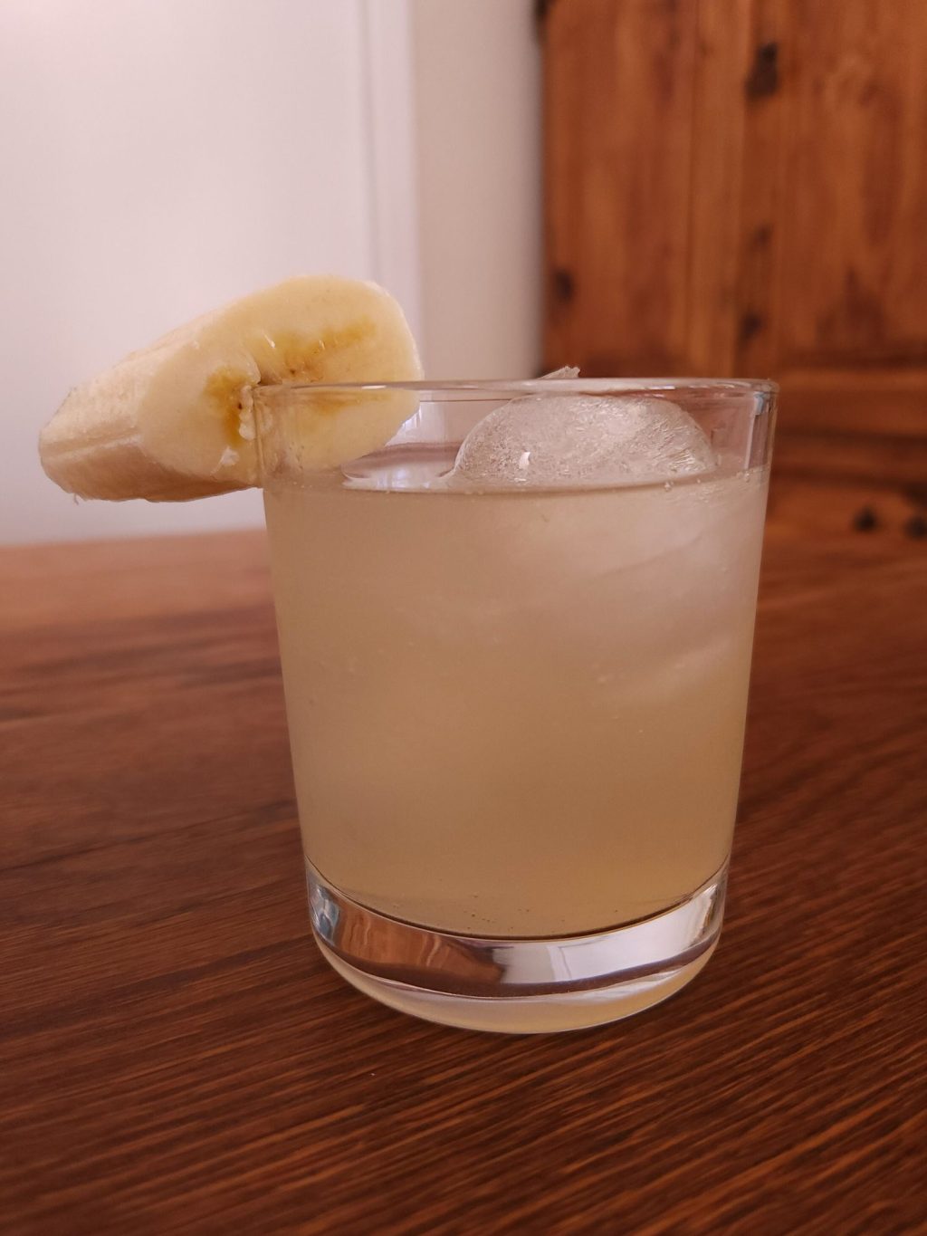Rocks glass with two ice spheres, light yellow liquid, and a banana slice garnish on the rim