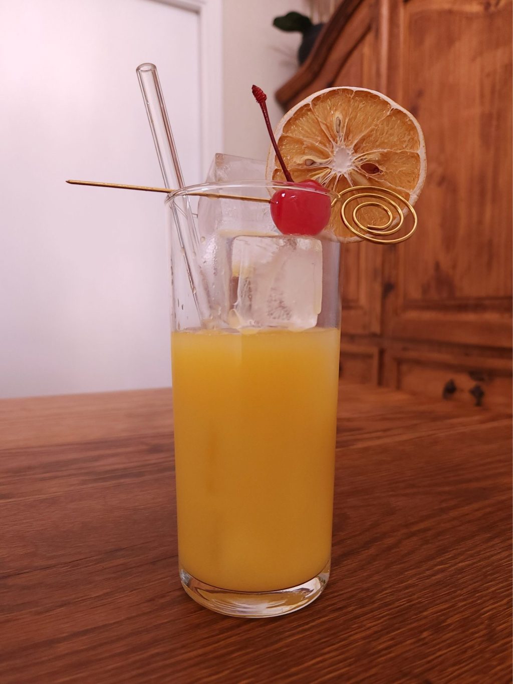 Tall glass with ice and bright orange yellow liquid, a clear glass straw, a bright red cherry perched on the rim with a rose gold cocktail pick, and a dried orange wheel garnish.