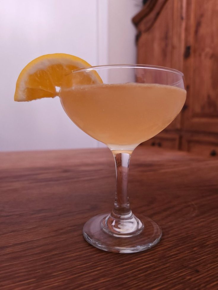 Coupe glass with yellow liquid and a lemon slice garnish, sitting on a wooden table