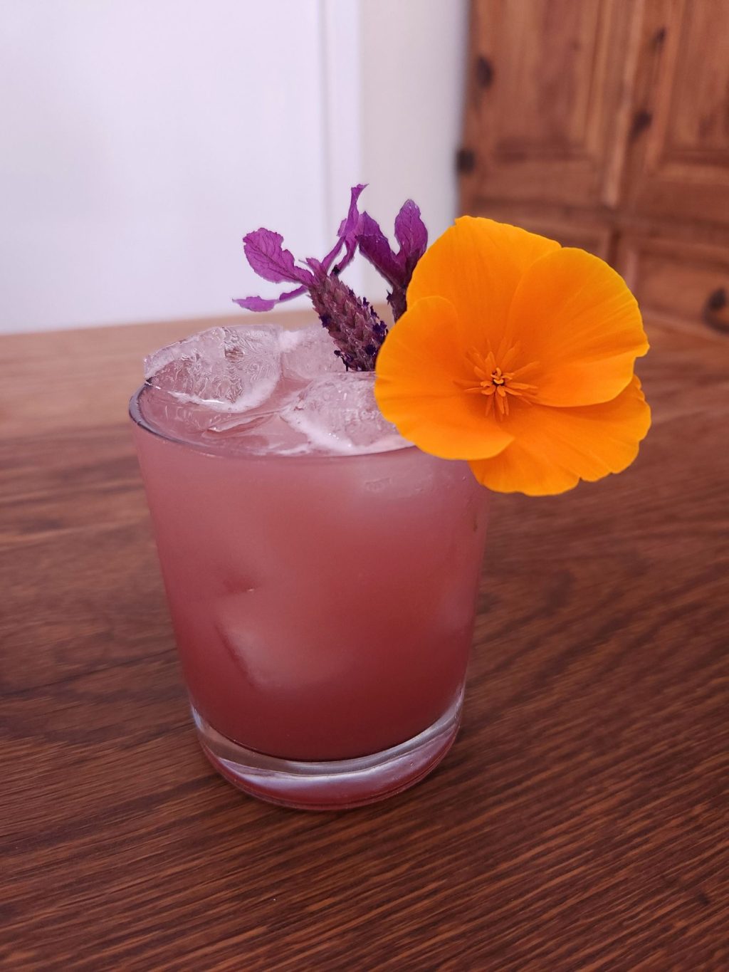 Short glass filled with ice and rosy purple liquid, with a garnish of lavender sprigs and a large California Poppy blossom