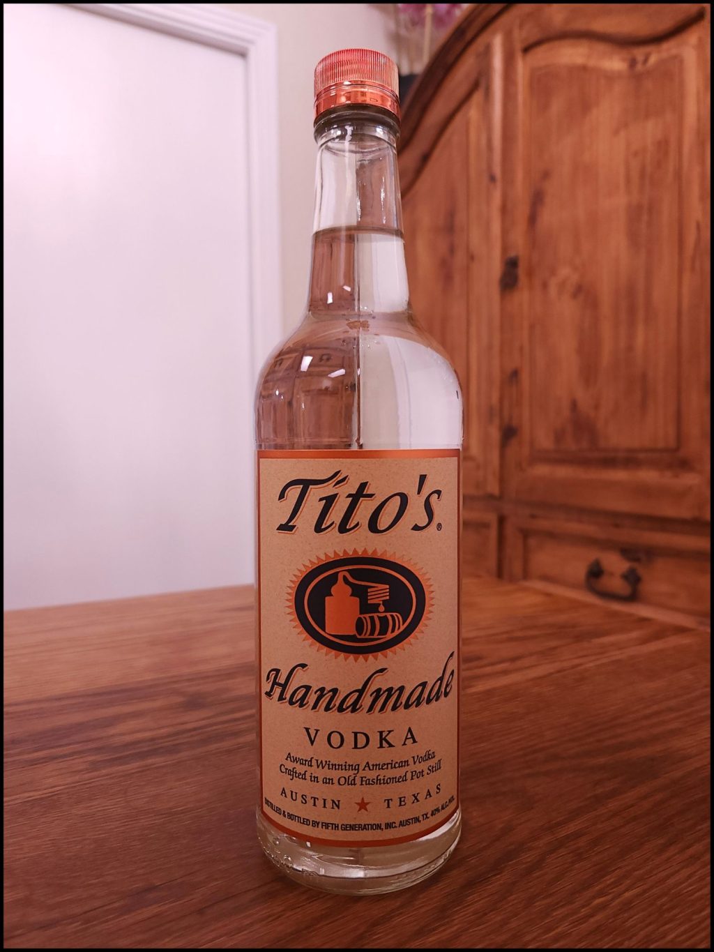 Bottle of Tito's Handmade Vodka with clear liquid and a light brown, black, and copper label design, sitting on a wooden table