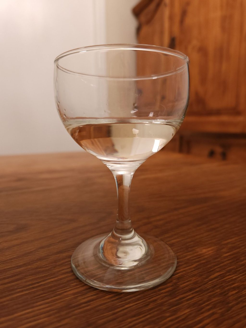 Small stemmed glass with lightly golden clear liquid, sitting on a wooden table